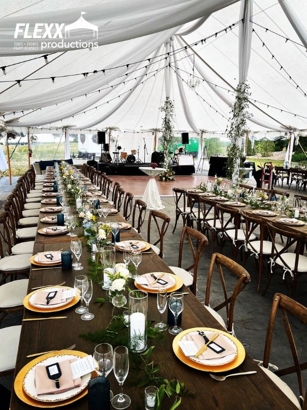 pole-tent-table-chair-wedding-rentals Long wooden tables and chairs, placesettings, market lighting, and a stage set up in a pole tent for a wedding reception