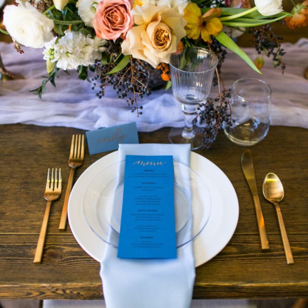 visit-our-wedding-rental-showroom A wedding reception place setting with flatware, a charger, dinnerplate, glassware, and a blue linen napkin